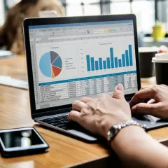 Osoba pracuje na laptopie, na ekranie widoczne są wykresy i dane. Widać tabelę z danymi, która może pomóc w nauce, jak obliczyć średnią arytmetyczną w Excelu.
