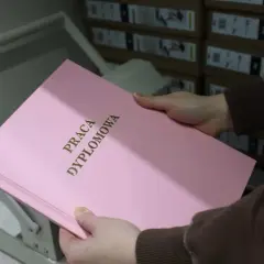 Różowa okładka pracy dyplomowej z napisem "PRACA DYPLOMOWA" w złoconych literach. W tle stosy pudeł, które mogą zawierać materiały do prezentacji.