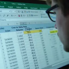 Osoba w okularach patrzy na arkusz kalkulacyjny Excel z danymi i formułami.