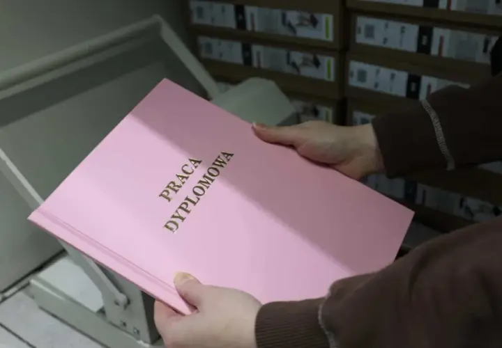 Różowa okładka pracy dyplomowej z napisem "PRACA DYPLOMOWA" w złoconych literach. W tle stosy pudeł, które mogą zawierać materiały do prezentacji.