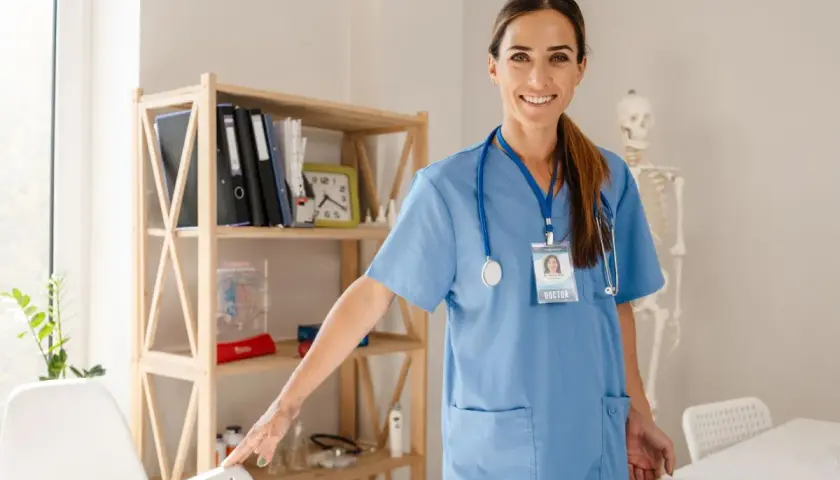 Lekarka w niebieskim kitlu z identyfikatorem "DOCTOR" uśmiecha się, stojąc obok regału z książkami i modelem szkieletu. Czy studia medyczne w Polsce są płatne?