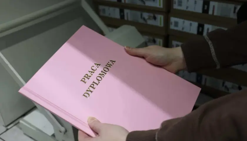 Różowa okładka pracy dyplomowej z napisem "PRACA DYPLOMOWA" w złoconych literach. W tle stosy pudeł, które mogą zawierać materiały do prezentacji.
