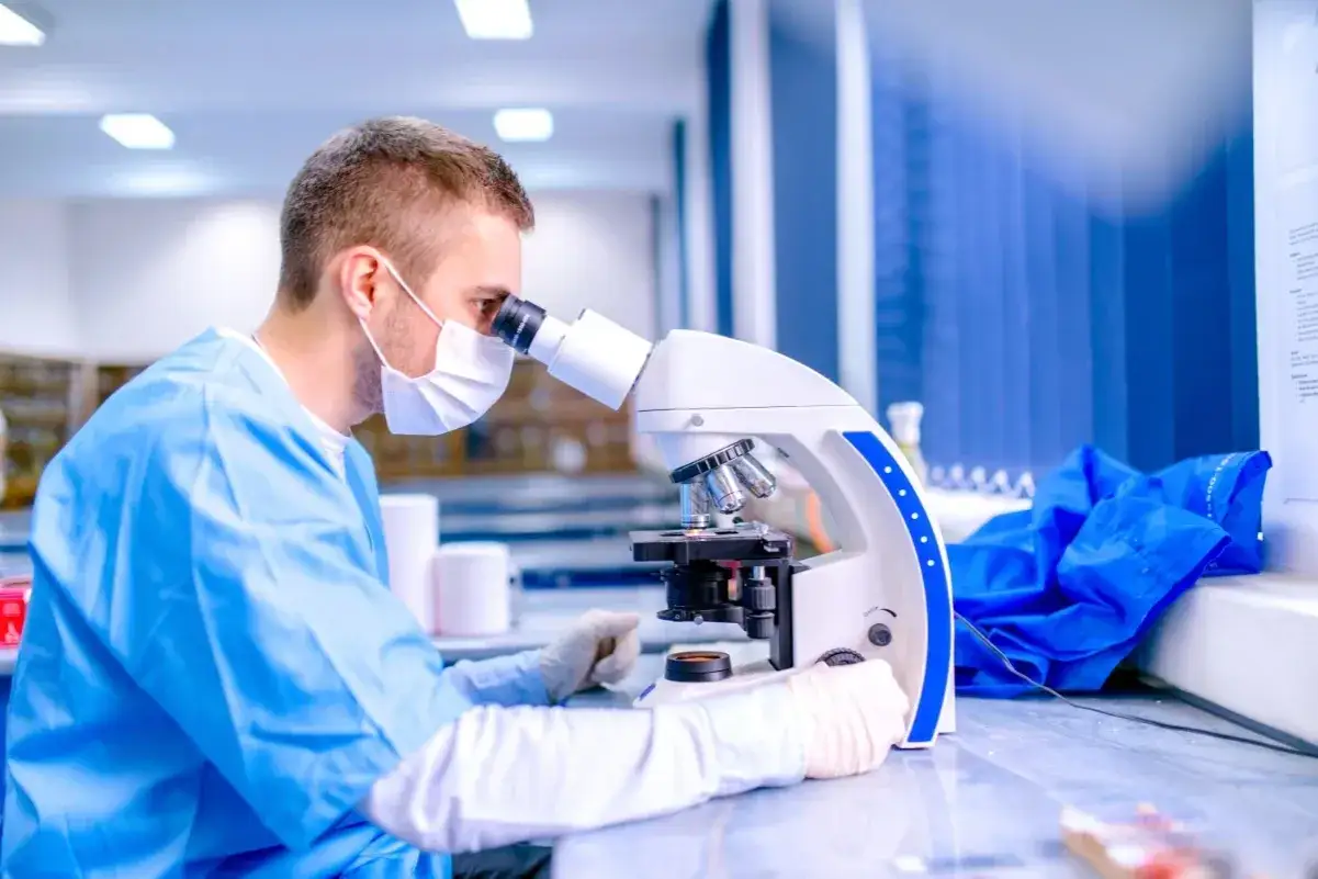 Mężczyzna w fartuchu i masce medycznej, kto to jest doktor nauk medycznych, bada próbki pod mikroskopem w laboratorium.