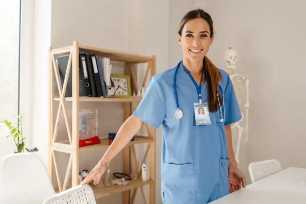 Lekarka w niebieskim kitlu z identyfikatorem "DOCTOR" uśmiecha się, stojąc obok regału z książkami i modelem szkieletu. Czy studia medyczne w Polsce są płatne?